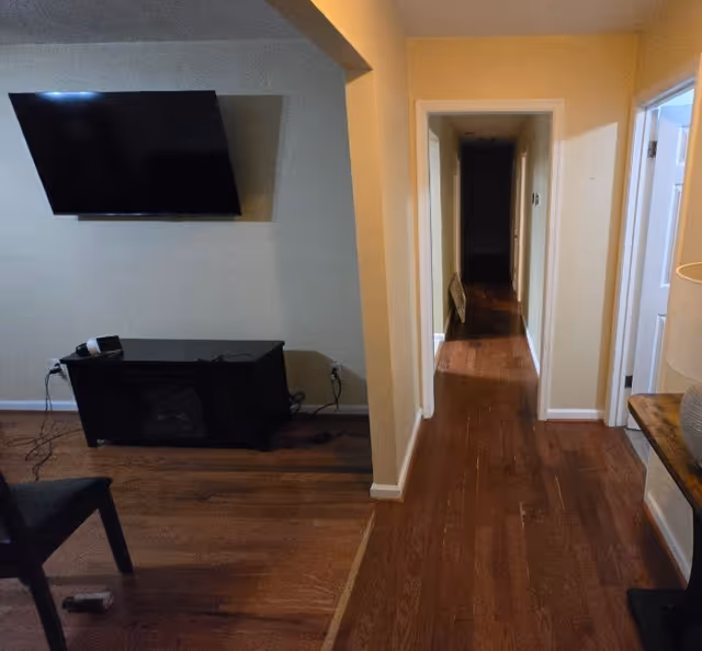 A living room with a wall-mounted TV and console beside a hallway leading to a dark corridor, all on hardwood floors.