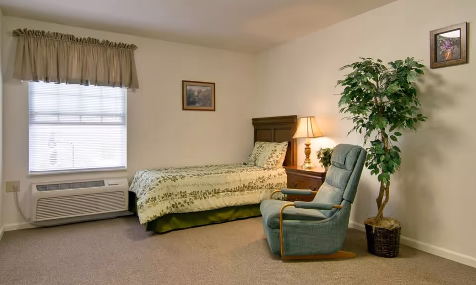 A simple bedroom with a single bed covered in a green and white floral bedspread, a wooden nightstand with a lamp, a green upholstered armchair, a potted artificial tree, two framed pictures on the walls, and a window with blinds and a beige valance.