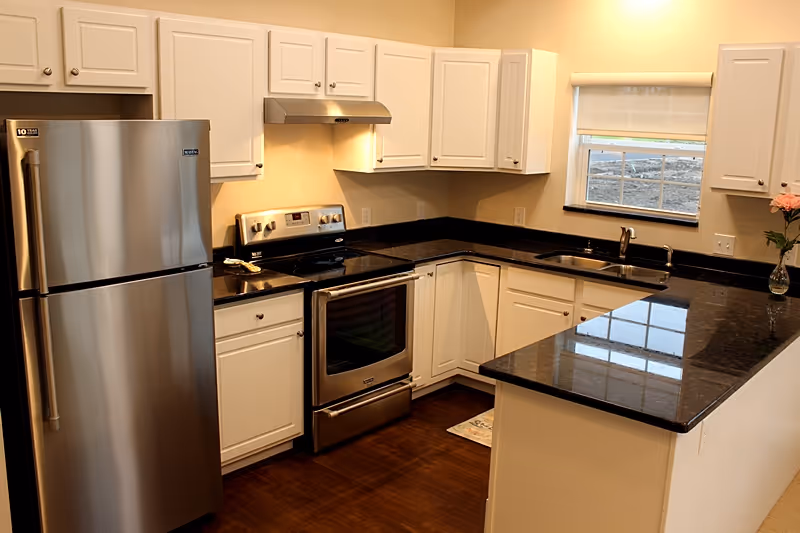 Modern kitchen with stainless steel refrigerator and stove, white cabinets, dark granite countertops, a sink, and a window.