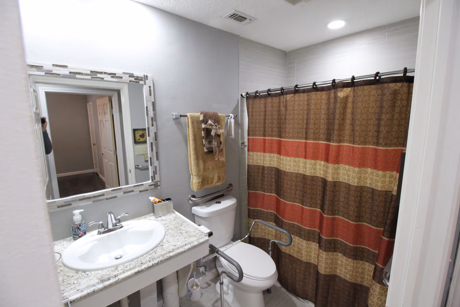 A bathroom with a white sink and granite countertop, a mirror above the sink, a toilet with safety grab bars on both sides, a towel rack with folded towels, and a shower with a brown, red, and beige striped curtain.