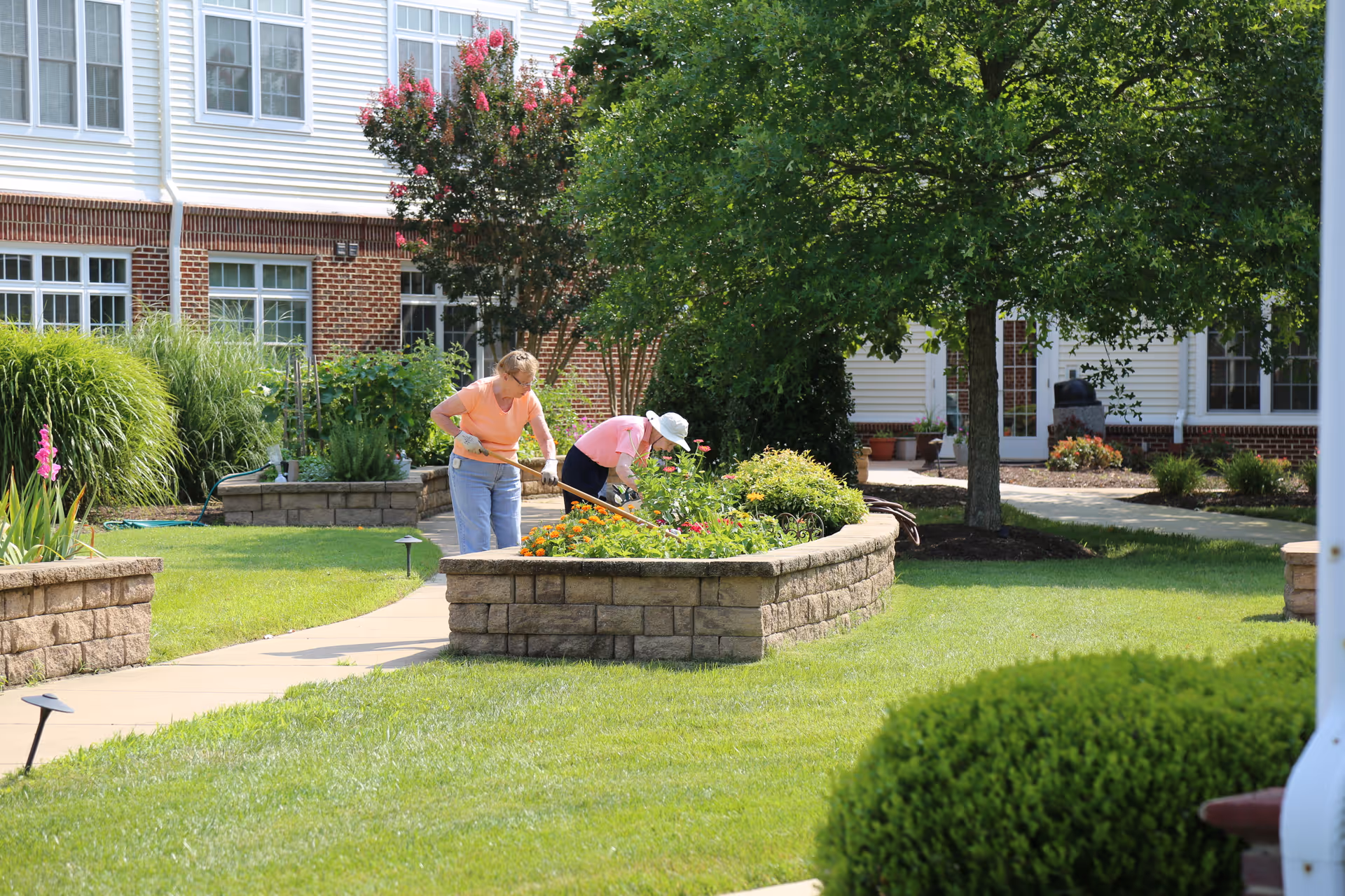 Two elderly women gardening in a raised flower bed in a well-maintained outdoor courtyard area of a senior living facility with green grass, trees, and a brick and white building in the background.