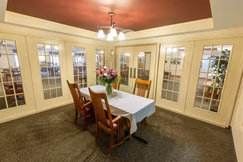 A small dining area with a rectangular table covered with a white tablecloth and four wooden chairs. A vase with a colorful bouquet of flowers is placed in the center of the table. The room has beige walls with multiple glass-paneled doors and a reddish ceiling with a modern light fixture.