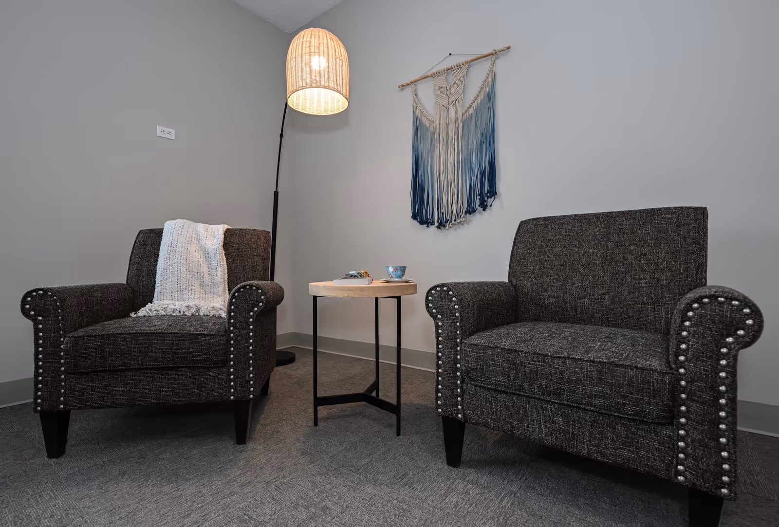 A cozy seating area with two dark gray armchairs featuring nailhead trim, a small round wooden side table between them holding a cup and a plate, a tall floor lamp with a woven lampshade, and a decorative blue and white macrame wall hanging on a light gray wall.