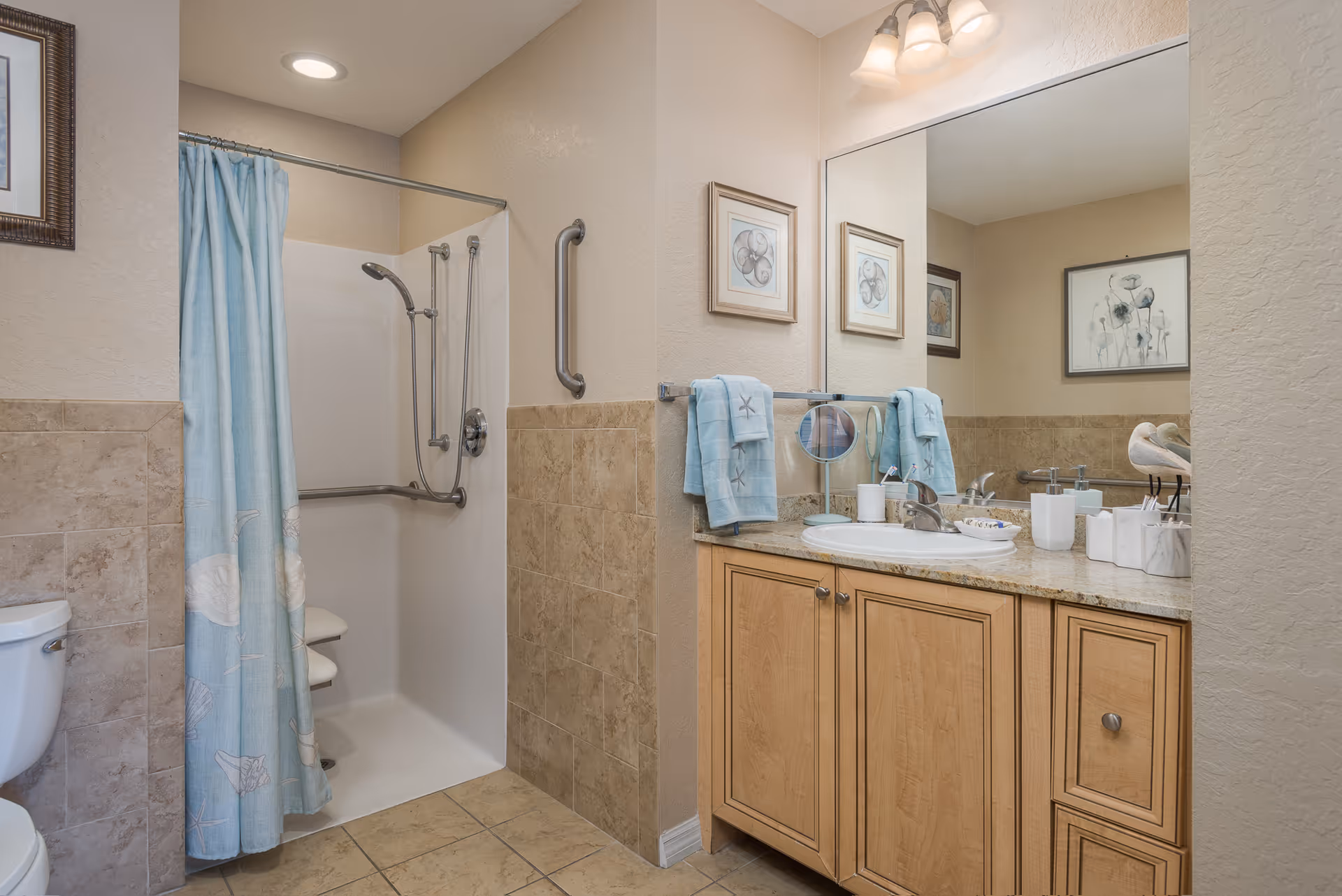 A bathroom with a walk-in shower featuring a blue shower curtain with seashell designs, a grab bar, and a fold-down shower seat. The bathroom also has a toilet, a wooden vanity with a granite countertop, a sink, and a large mirror above the vanity. Two blue towels hang on a towel rack, and there are framed pictures on the walls.