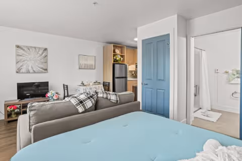Studio-style interior showing a bed in the foreground, a sofa and TV, a small dining table, kitchenette, and an open bathroom doorway.
