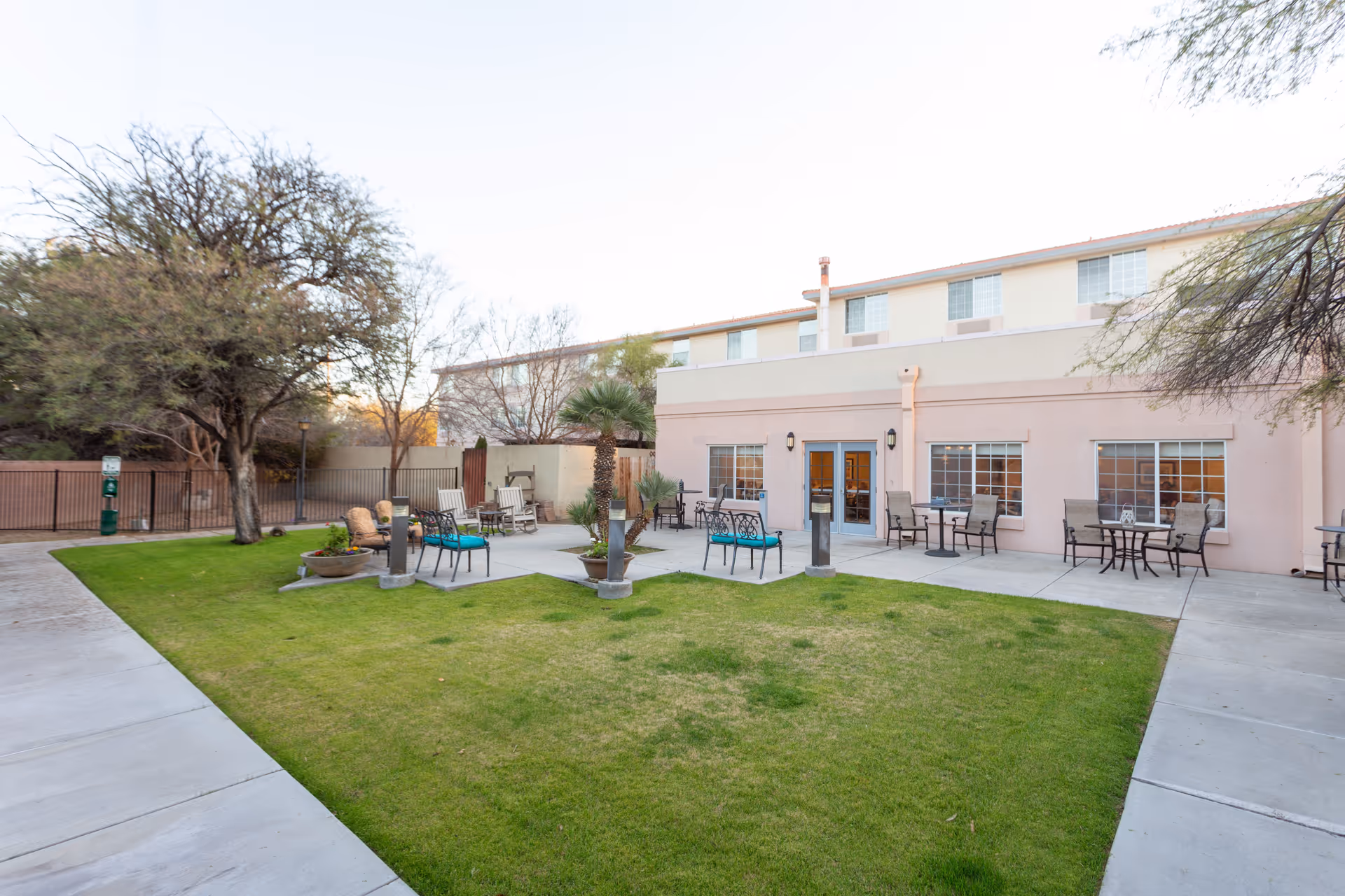 Outdoor patio area at Avista Senior Living Tucson featuring green lawn, several seating arrangements with chairs and tables, a few potted plants, and a two-story building with multiple windows in the background.