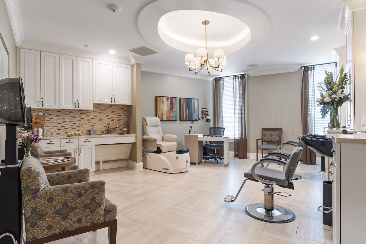 A bright and spacious salon area in a senior living facility featuring a pedicure chair, a manicure table with a desk lamp, two salon styling chairs, and a washing station. The room has light-colored tiled flooring, white cabinetry with a mosaic tile backsplash, two large windows with brown curtains, and a modern chandelier hanging from a recessed ceiling. There are also two patterned armchairs and a small table with flowers and magazines.
