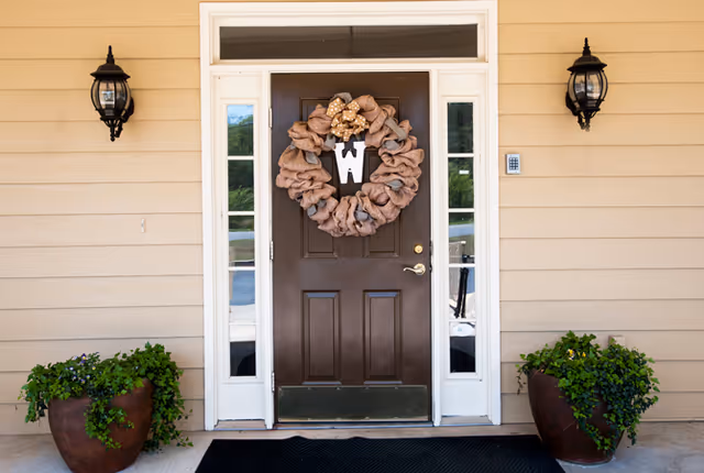 Front entrance with a brown door decorated with a wreath, sidelights, wall lanterns, and potted plants.