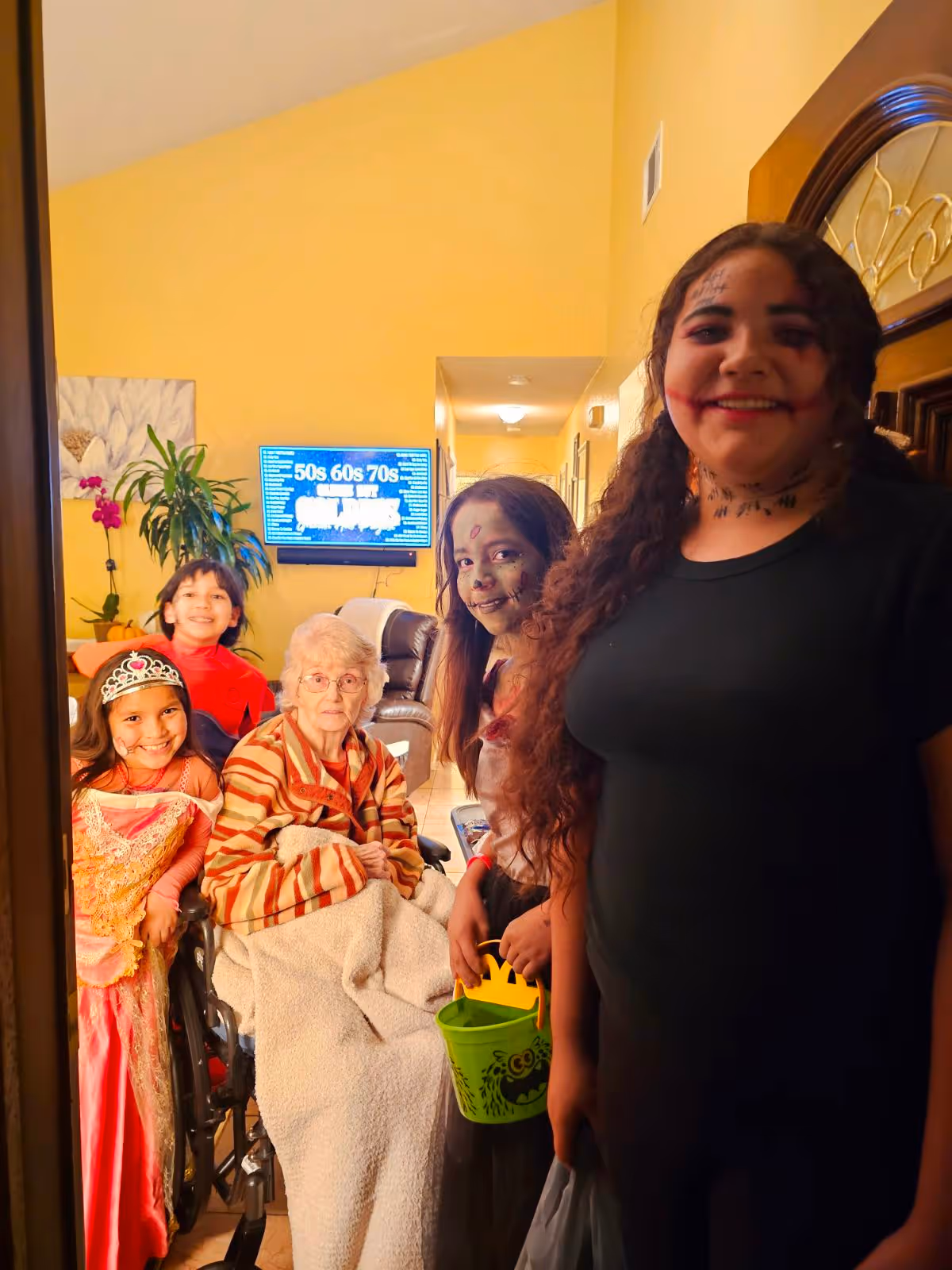 An elderly woman in a wheelchair is surrounded by four children dressed in Halloween costumes inside a warmly lit room. The children are smiling and one holds a green bucket with a Halloween design. The room has yellow walls, a TV mounted on the wall displaying text, a plant, and a painting of a flower.