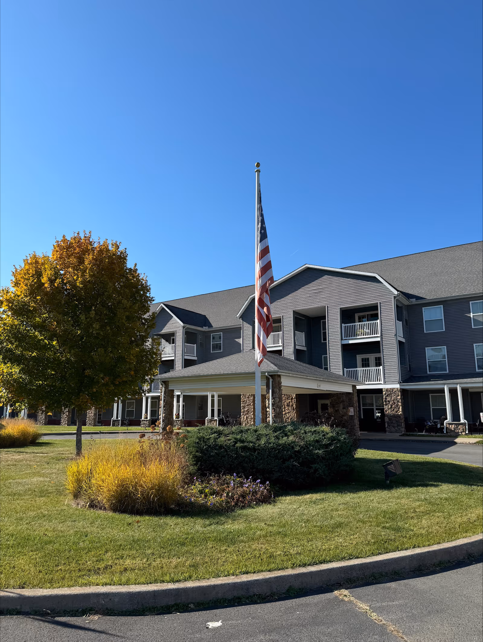 Front entrance of a multi-story senior living building with an American flag on a flagpole, landscaped lawn, and balconies under a clear blue sky.