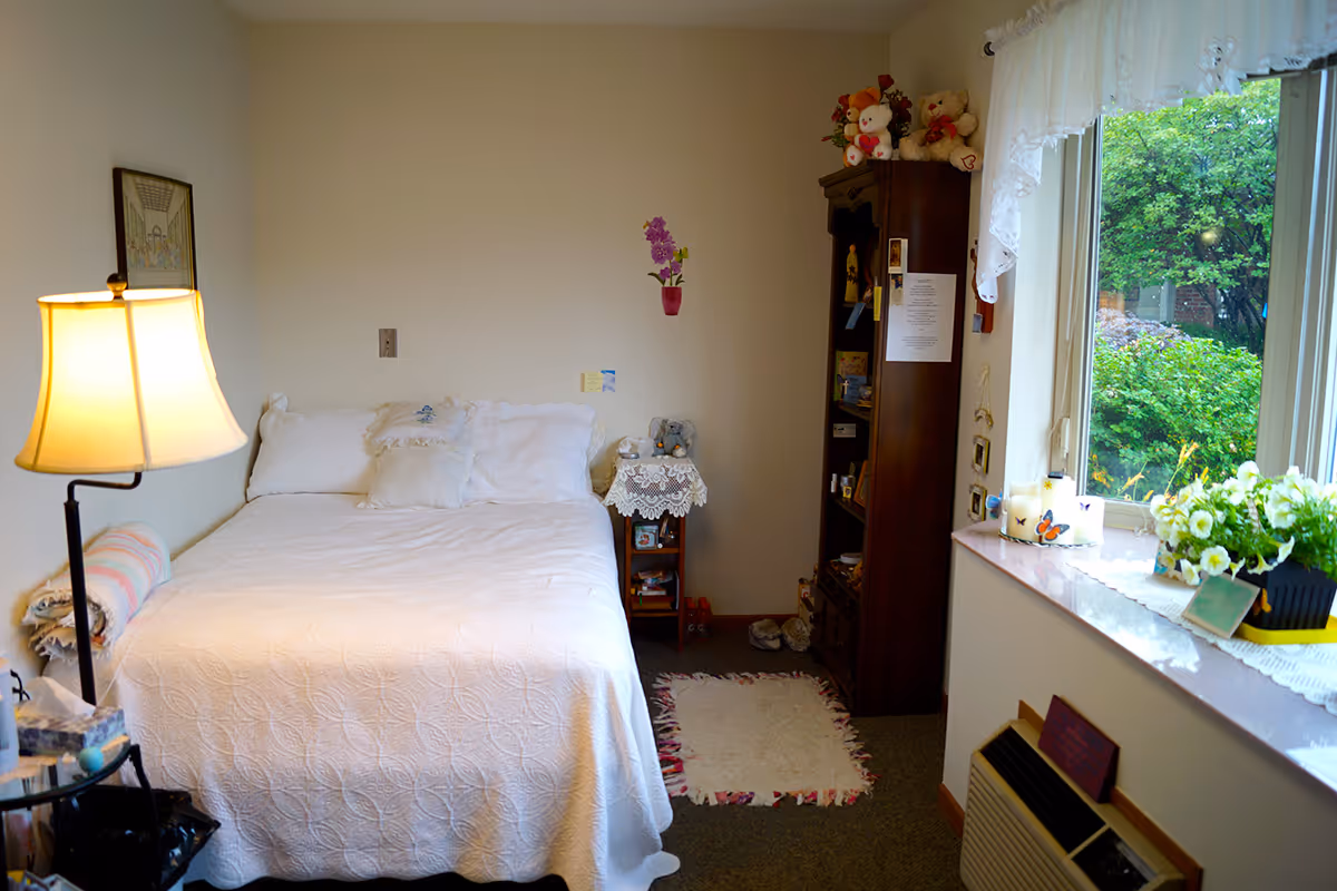 Cozy assisted-living bedroom with a neatly made bed, floor lamp, wooden shelving, and a window with plants looking out to greenery.