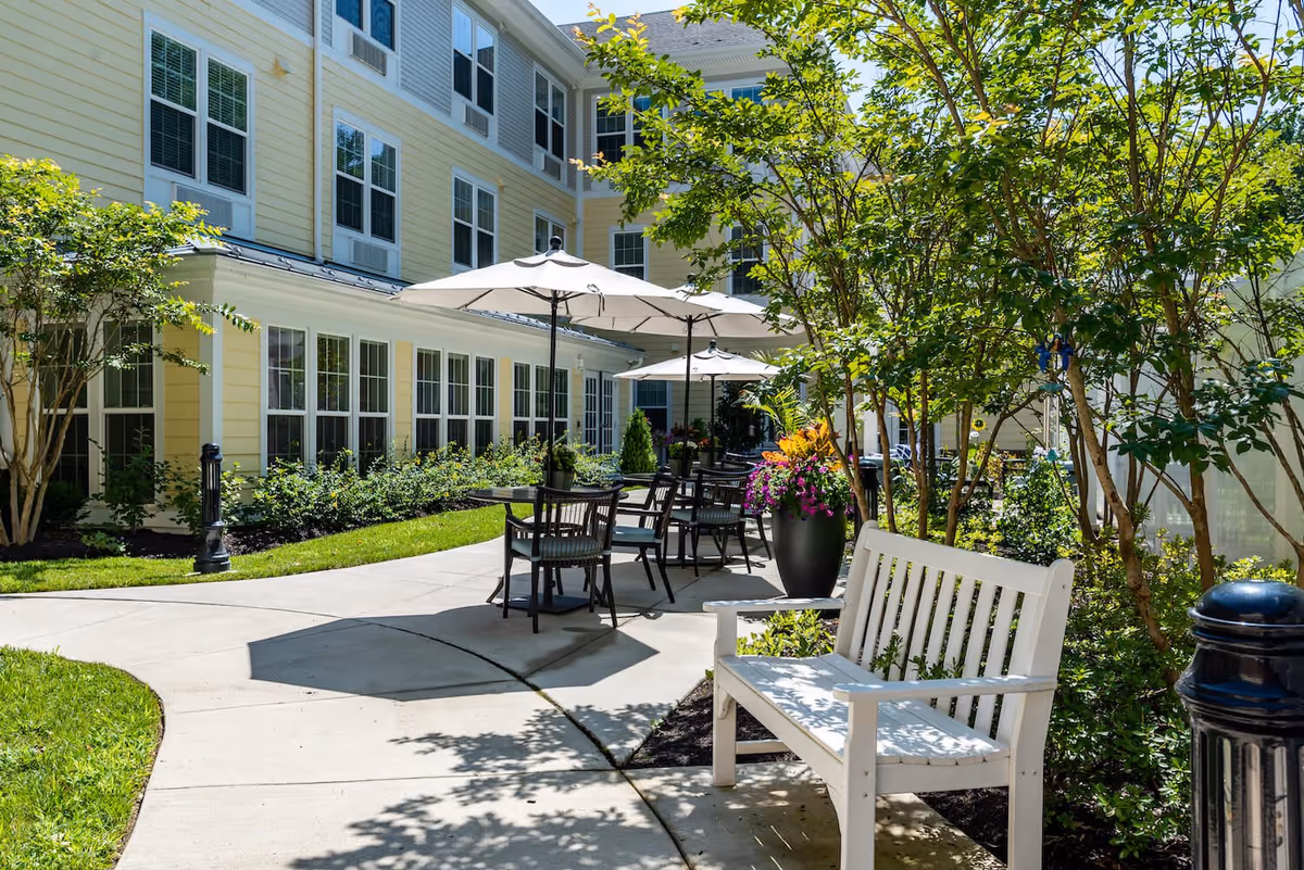 Outdoor patio area at Arbor Terrace Waugh Chapel featuring a curved concrete walkway, white wooden bench, several tables with chairs and large umbrellas, surrounded by greenery and flowering plants next to a yellow building with many windows.