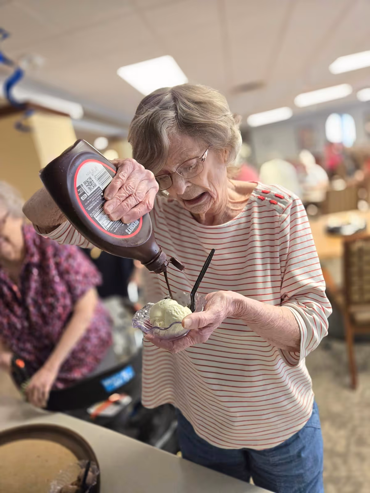 An elderly woman wearing glasses and a striped shirt is pouring chocolate syrup onto a scoop of ice cream in a small dish, with a black spoon in the dish. Another elderly woman is visible in the background in a communal dining area.