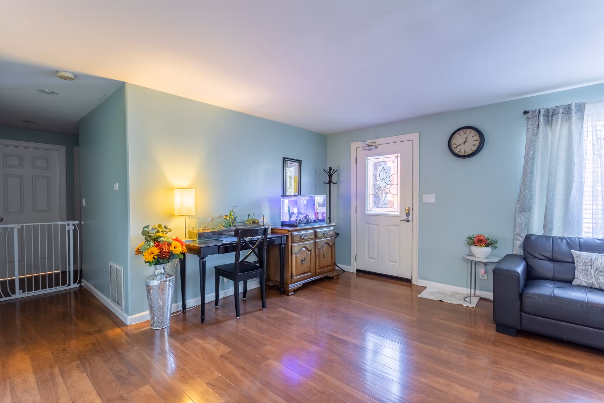 A bright living room with light blue walls and wooden flooring. There is a black leather couch with a pillow on the right, a small round side table with a flower arrangement, and a window with sheer curtains. Near the center, there is a wooden cabinet with a fish tank on top, a black chair, and a small desk with a lamp and flowers. A white door with decorative glass is visible, along with a wall clock above it. A baby gate is installed in the hallway on the left side.