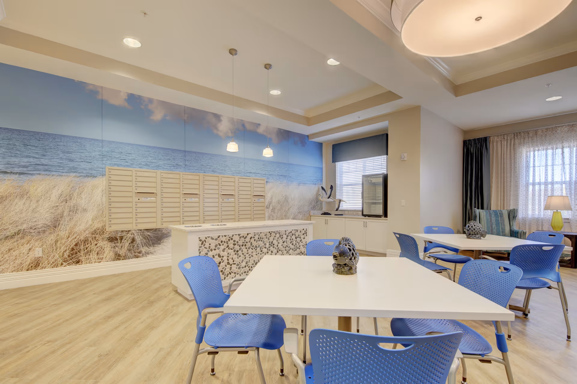 A bright and spacious common area with white tables and blue chairs. The back wall features a large mural of a beach scene with sand dunes and ocean waves. There are mailboxes mounted on the wall beneath the mural. The room has wood flooring, pendant lights hanging from the ceiling, and a window with blinds and curtains. A small seating area with a lamp and cushions is visible in the background.