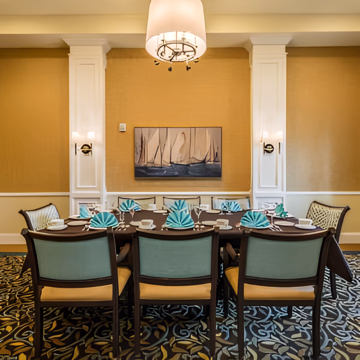 A formal dining room with a rectangular table set for eight people. The table is covered with a dark tablecloth and has turquoise folded napkins, white plates, and glassware. The room features beige walls with white trim and columns, a patterned carpet, a modern chandelier hanging above the table, and a painting of sailboats on the wall.