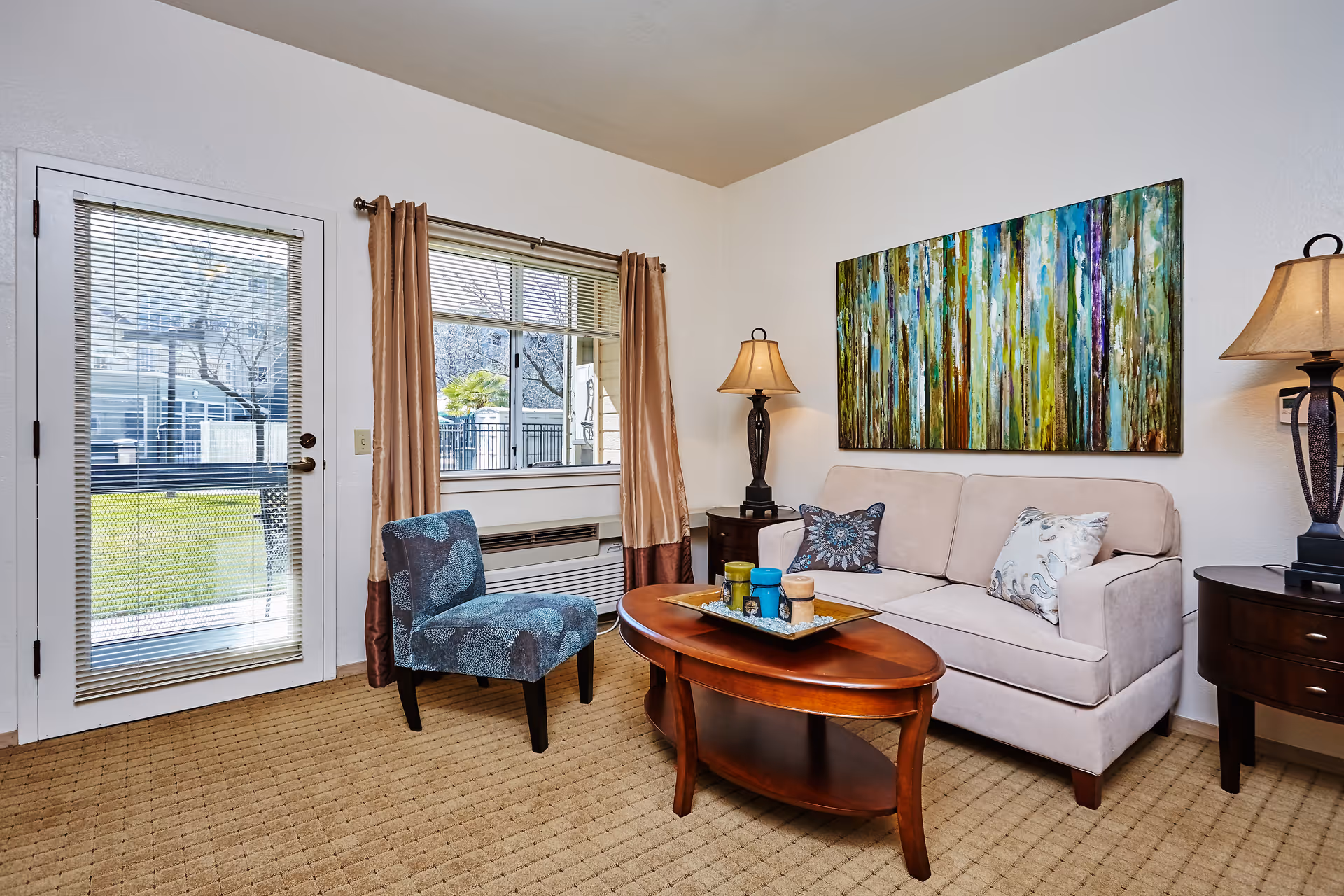 A cozy living room with a beige sofa adorned with decorative pillows, a wooden oval coffee table with candles on a tray, a blue patterned armchair, two dark wooden side tables each with a lamp, a large colorful abstract painting on the wall, a window with beige curtains, and a glass door leading outside.