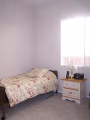 A small bedroom with a single bed covered in a floral patterned comforter. Next to the bed is a wooden nightstand with two drawers, a table lamp, and a small radio or clock. A window with blinds is on the wall behind the nightstand, allowing natural light into the room.