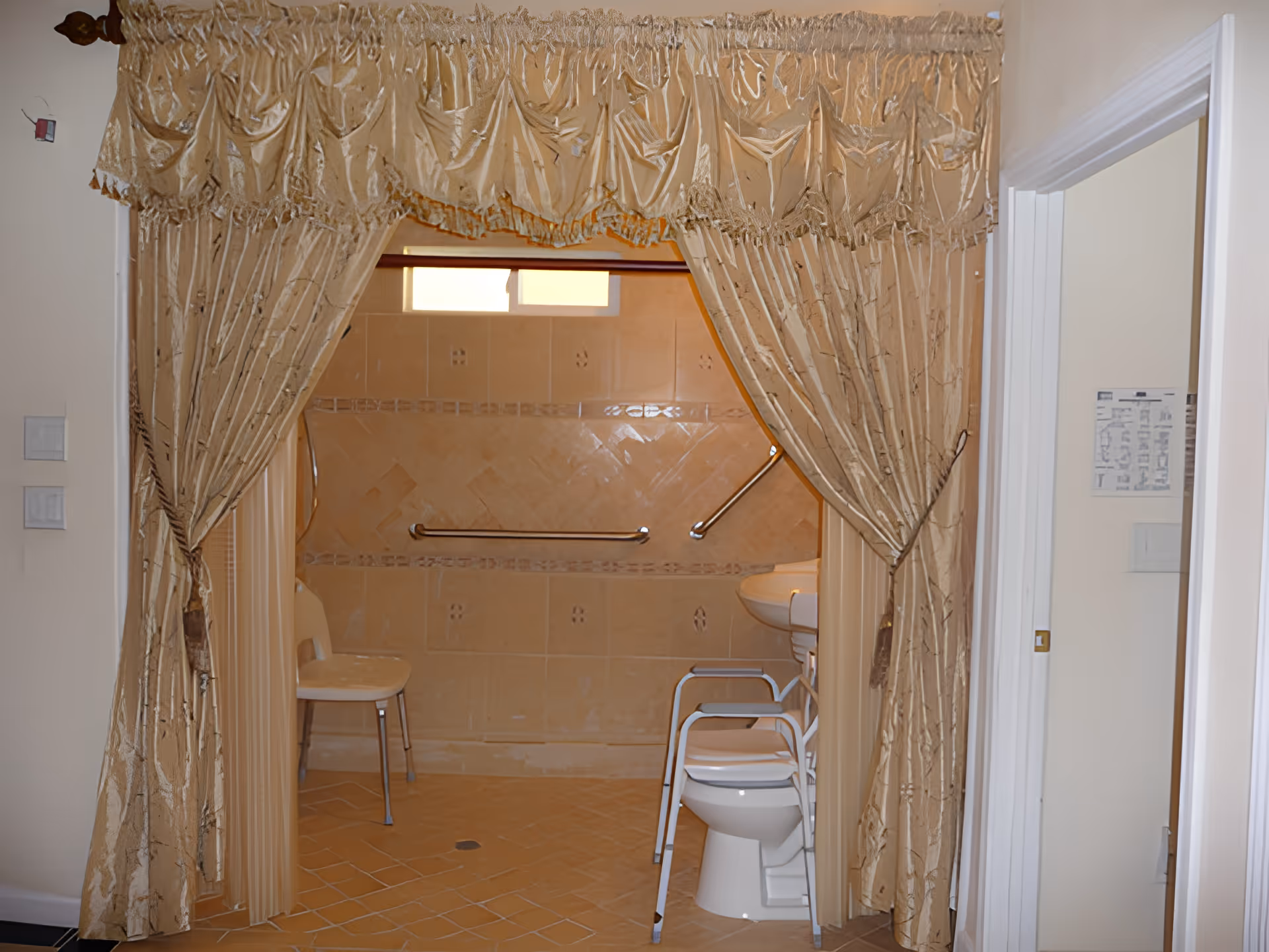 View through a doorway with gold-colored curtains tied back, revealing a bathroom with beige tiled walls and floor. Inside the bathroom, there is a white chair, a toilet with a raised seat and armrests, a sink, and grab bars on the walls for support.