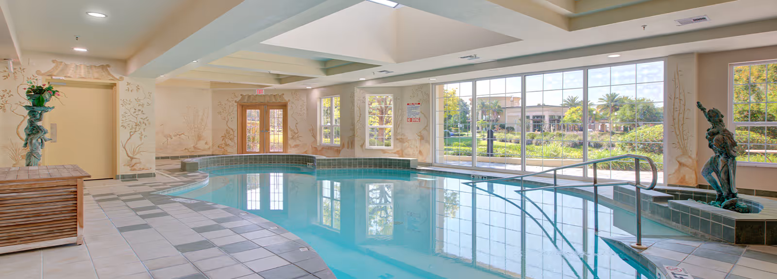 Indoor swimming pool area with tiled floor and walls featuring decorative murals. Large windows provide a view of a garden with palm trees and buildings outside. The pool has a handrail for easy access and two statues on either side of the pool area.