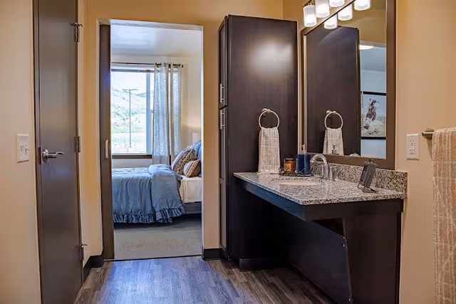 View of a bathroom vanity with a granite countertop, dark wood cabinets, and a large mirror. A towel hangs on a ring next to the sink. Through an open doorway, a bedroom with a bed covered in blue bedding and a window with curtains is visible.