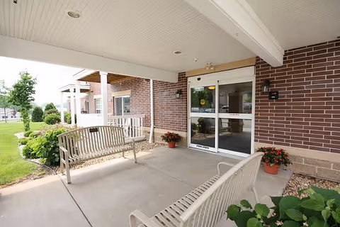 Covered entrance area of a senior living facility with two metal benches facing each other, potted plants on either side of the automatic sliding glass doors, and a brick exterior wall. There is a well-maintained lawn and shrubs visible to the left.