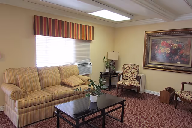 A cozy living room with a striped sofa, two floral-patterned armchairs, a black coffee table with a potted plant, a side table with a lamp and another plant, a large floral painting on the wall, and a window with striped valance and blinds. The room has beige walls and a patterned carpet.