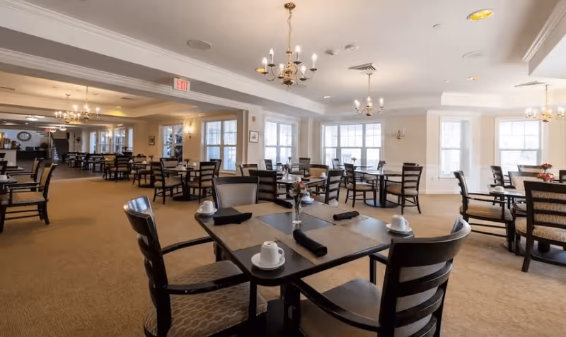 A spacious dining room with multiple tables and chairs arranged neatly. Each table is set with placemats, cups, and black napkins. The room is well-lit with large windows allowing natural light and several chandeliers hanging from the ceiling. The carpeted floor and light-colored walls create a warm and inviting atmosphere.