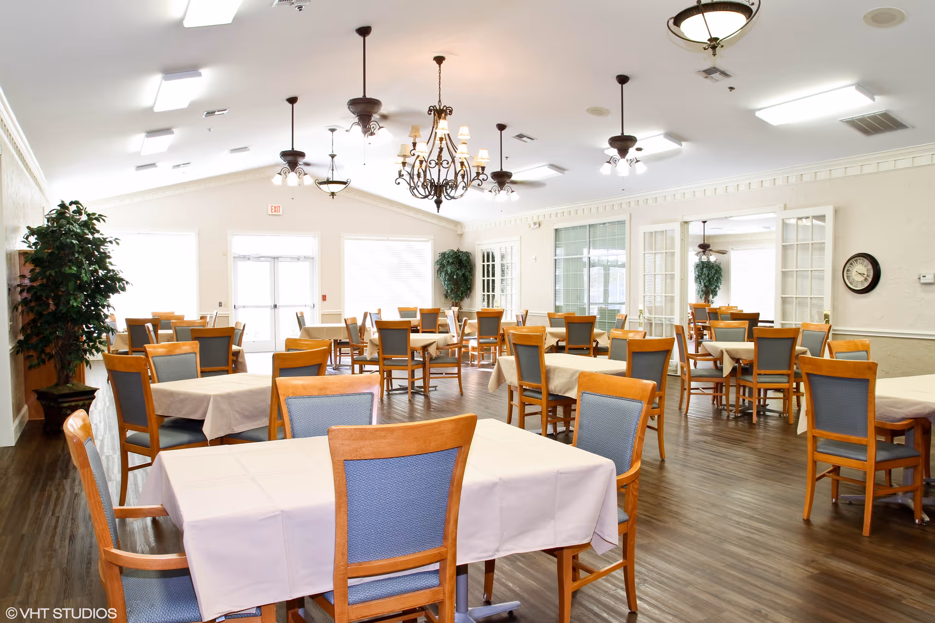 A bright and spacious dining room with multiple wooden tables covered with beige tablecloths and surrounded by wooden chairs with blue cushions. The room features several ceiling fans and chandeliers, large windows letting in natural light, potted plants, and a wall clock. The floor is dark wood, and the walls are light-colored with decorative molding.
