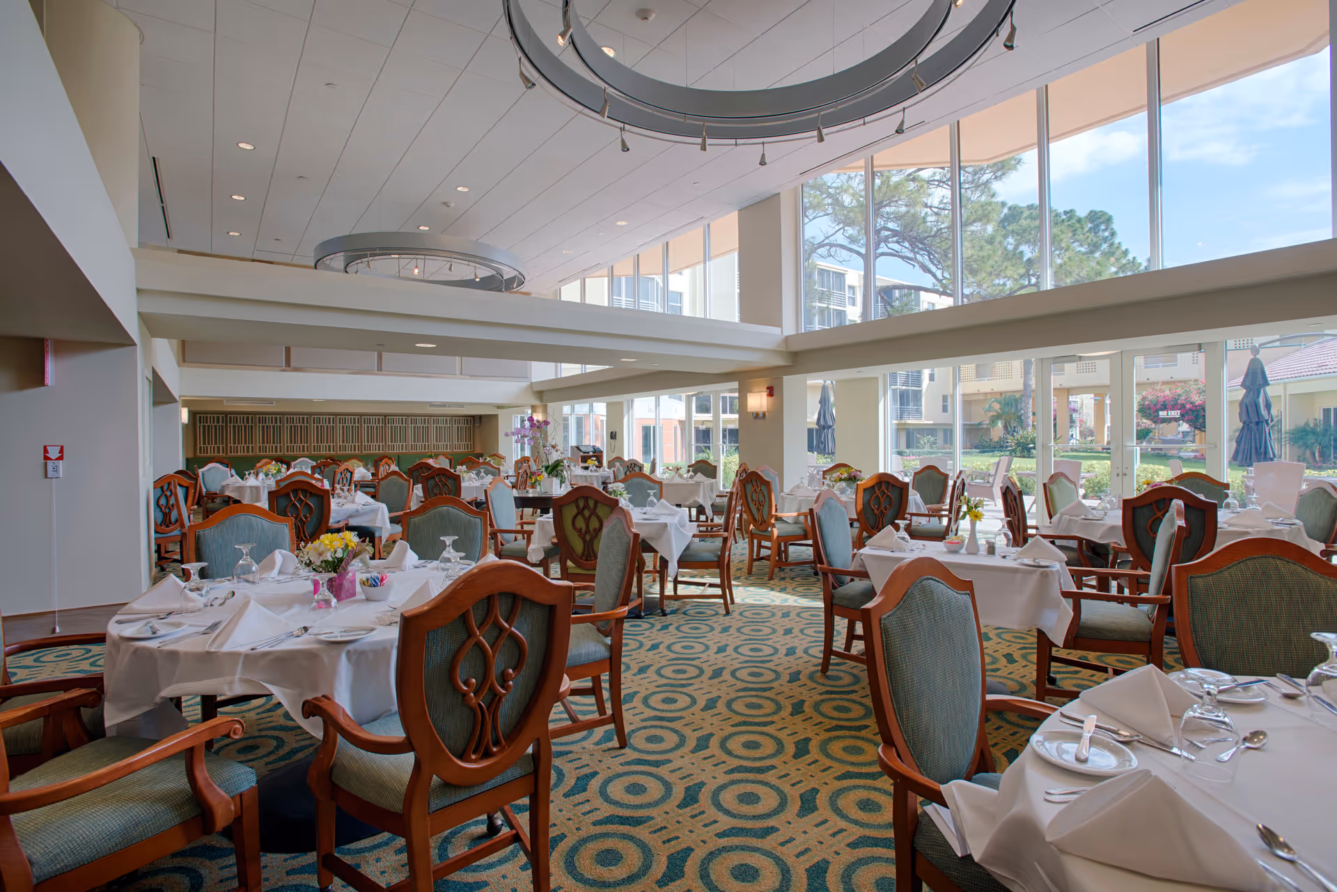 A bright and spacious dining room with large floor-to-ceiling windows allowing natural light to fill the space. The room is furnished with multiple round tables covered with white tablecloths, set with plates, glasses, and silverware. Each table is surrounded by wooden chairs with green upholstered seats and backs. The carpet has a geometric pattern in shades of green and beige. Outside the windows, there is a view of a garden and building exterior.