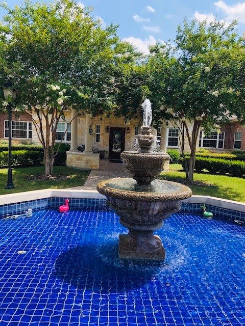 Outdoor view of a courtyard at Cypress Assisted Living - CyFair featuring a multi-tiered stone fountain in the center of a blue-tiled pool with small decorative flamingos. The courtyard is surrounded by green trees, manicured bushes, and a brick building with windows and a covered entrance.