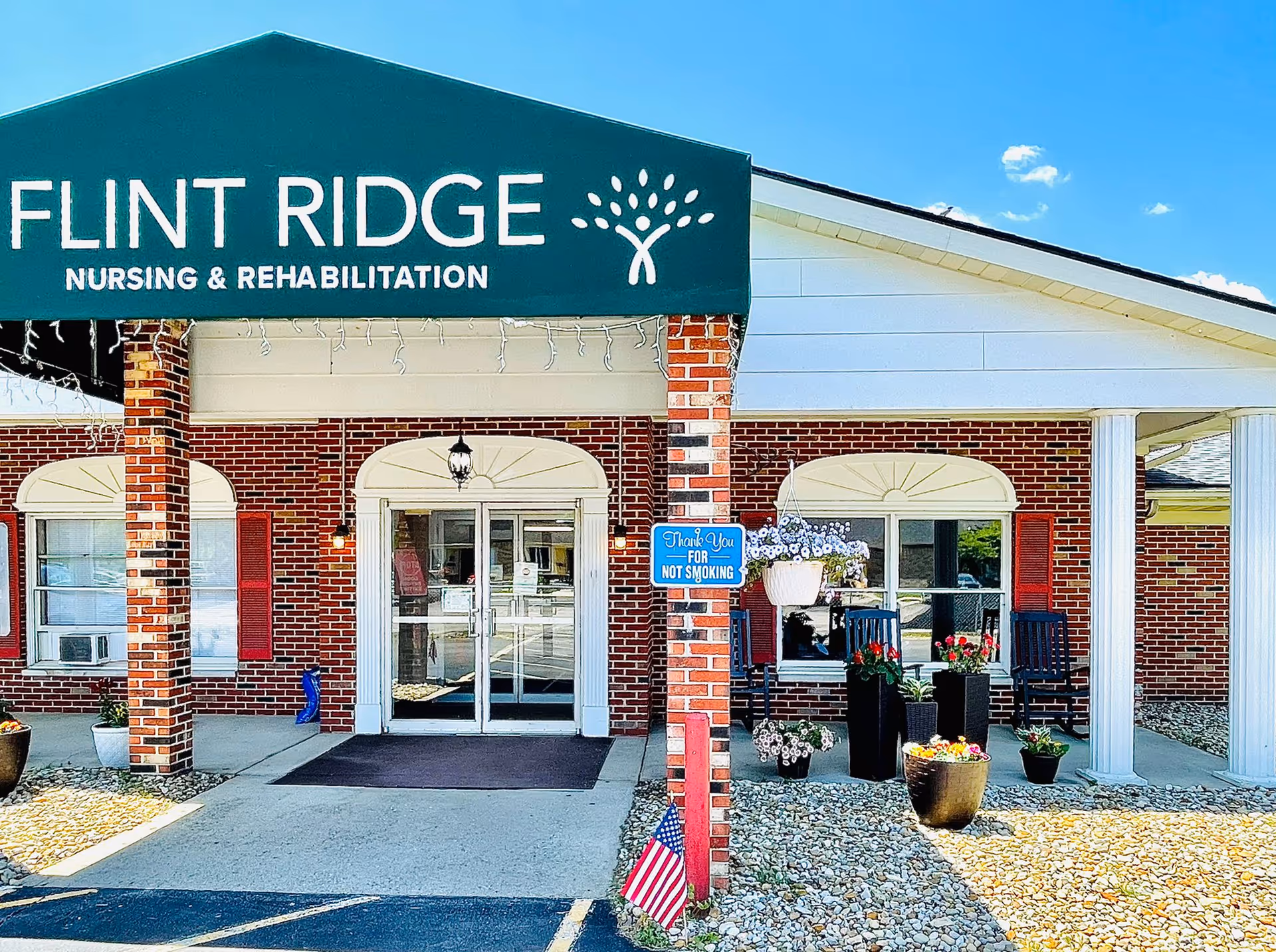Entrance of Flint Ridge Nursing and Rehabilitation facility with a green awning displaying the facility name. The building has red brick walls, white columns, and a glass double door entrance. There are potted plants and flowers near the entrance, a small American flag planted in the ground, and a blue sign that says 'Thank You For Not Smoking'.