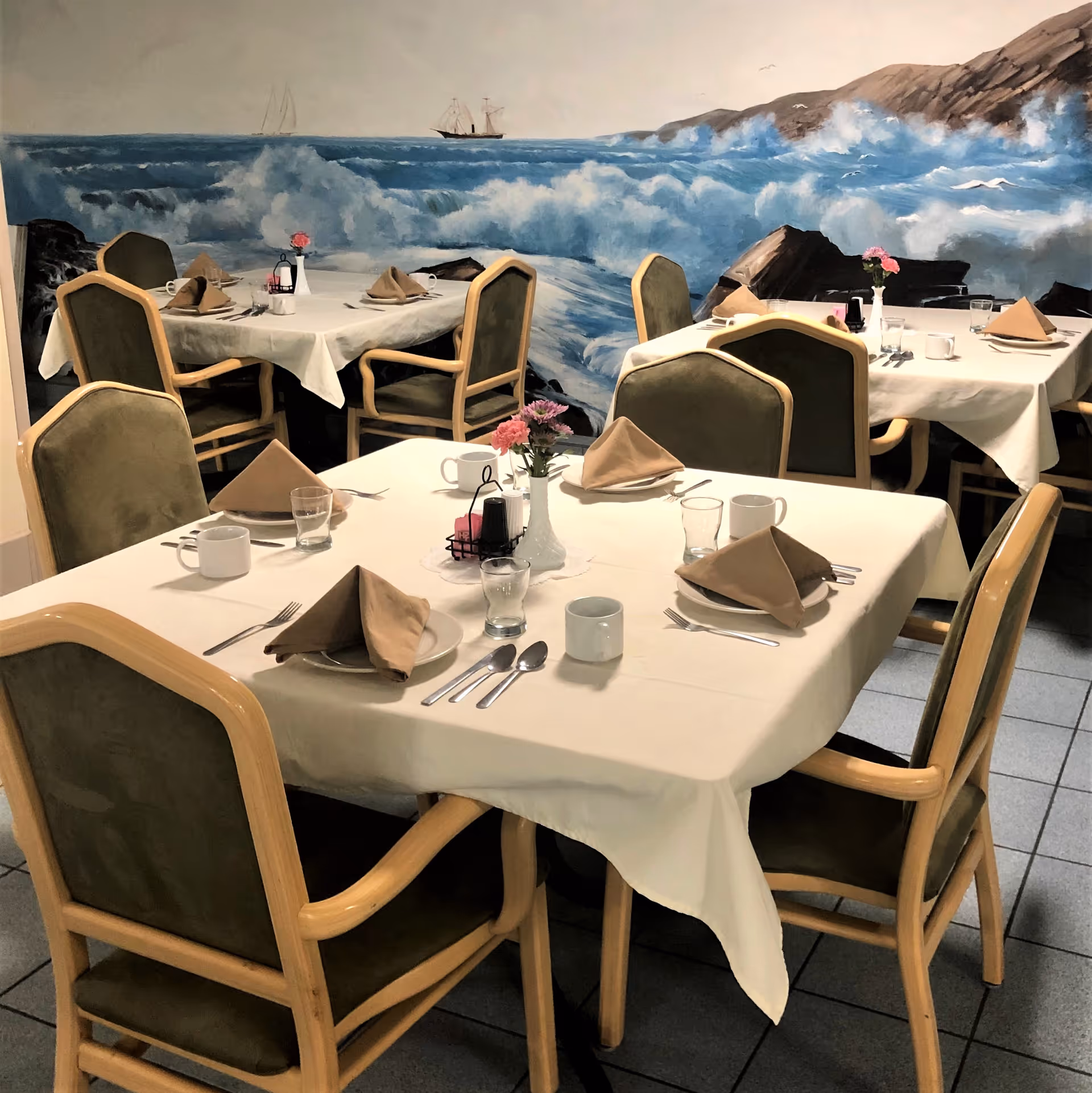 Dining area with tables covered in white tablecloths, each set with brown folded napkins, plates, cups, glasses, and silverware. The chairs have wooden frames with green upholstery. A mural of ocean waves crashing against rocks and a sailing ship is painted on the wall in the background.