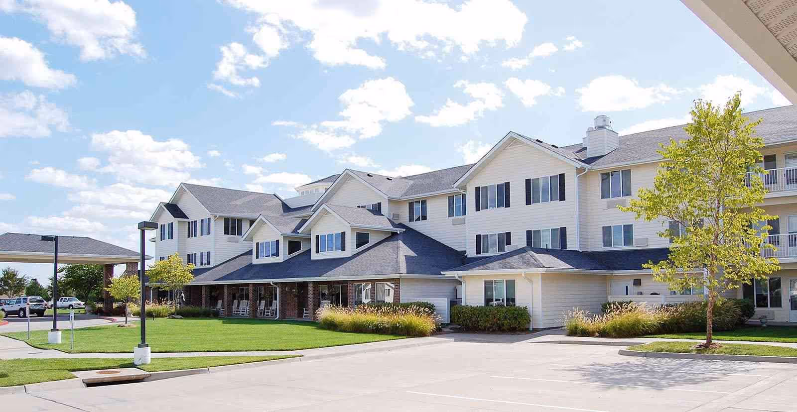 Exterior view of a large, multi-story senior living facility building with white siding and black shutters under a partly cloudy blue sky. The building has a covered entrance with rocking chairs and is surrounded by a well-maintained lawn, trees, and a paved driveway with parked cars visible in the background.