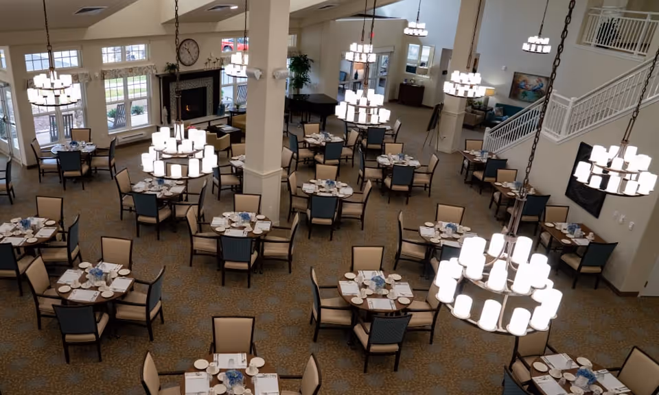 A spacious dining room with multiple round tables set with plates, napkins, and glasses. The room features beige and blue chairs, large chandeliers hanging from the ceiling, a fireplace with a clock above it, and a staircase leading to an upper level. Large windows allow natural light to enter the room.