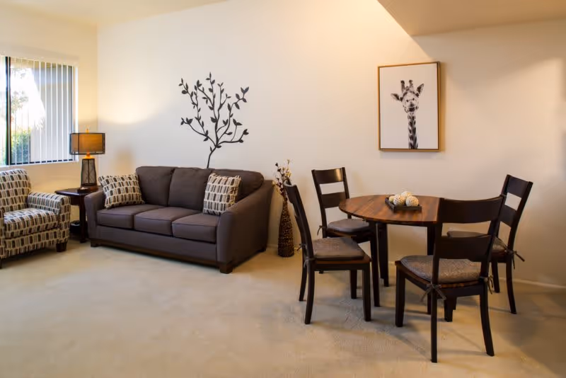 A cozy living area featuring a dark gray sofa with patterned cushions, two matching armchairs, a wooden side table with a lamp, a round wooden dining table with four chairs, a wall decal of a tree, and a framed picture of a giraffe on the wall.