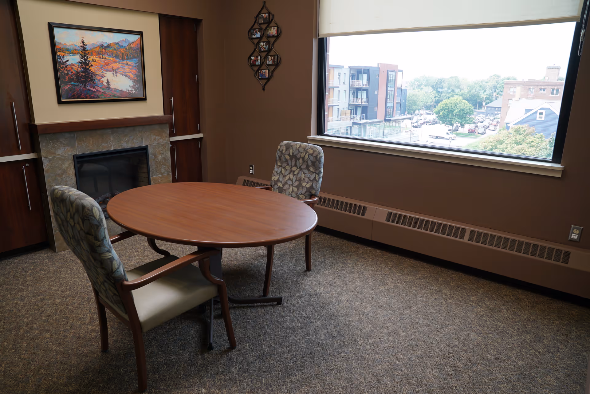 Small sitting room with a round wooden table, two upholstered chairs, a fireplace, wall art, and a large window overlooking nearby buildings.
