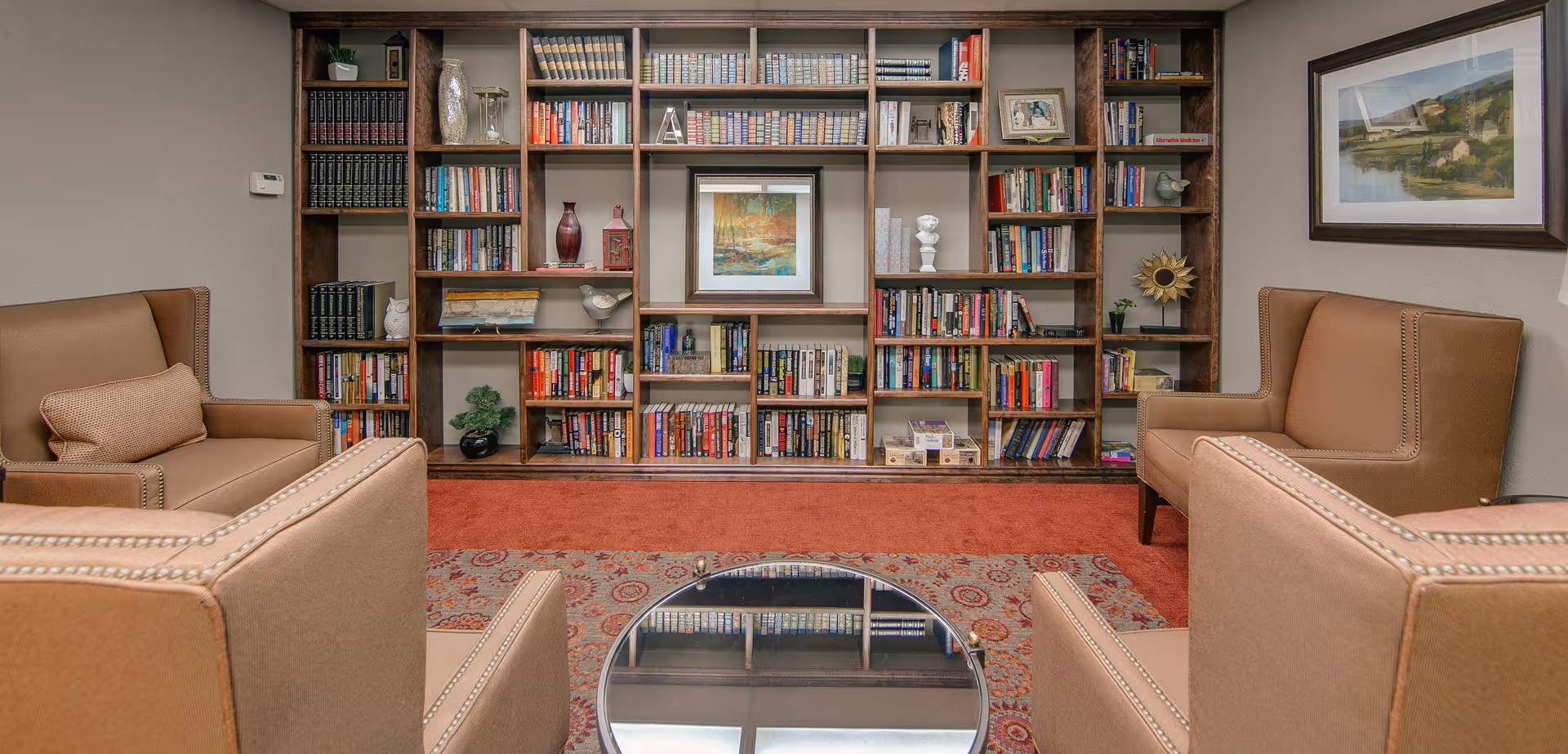 A cozy reading room with four tan armchairs arranged around a circular glass coffee table. Behind the chairs is a large wooden bookshelf filled with books and decorative items. The room has a red patterned carpet and a framed landscape painting on the wall to the right.