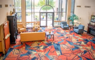 Bright and spacious senior living facility common area with large floor-to-ceiling windows letting in natural light. The room features colorful patterned carpet, a yellow sofa, several armchairs, a coffee table, and decorative plants. Outside greenery is visible through the windows.