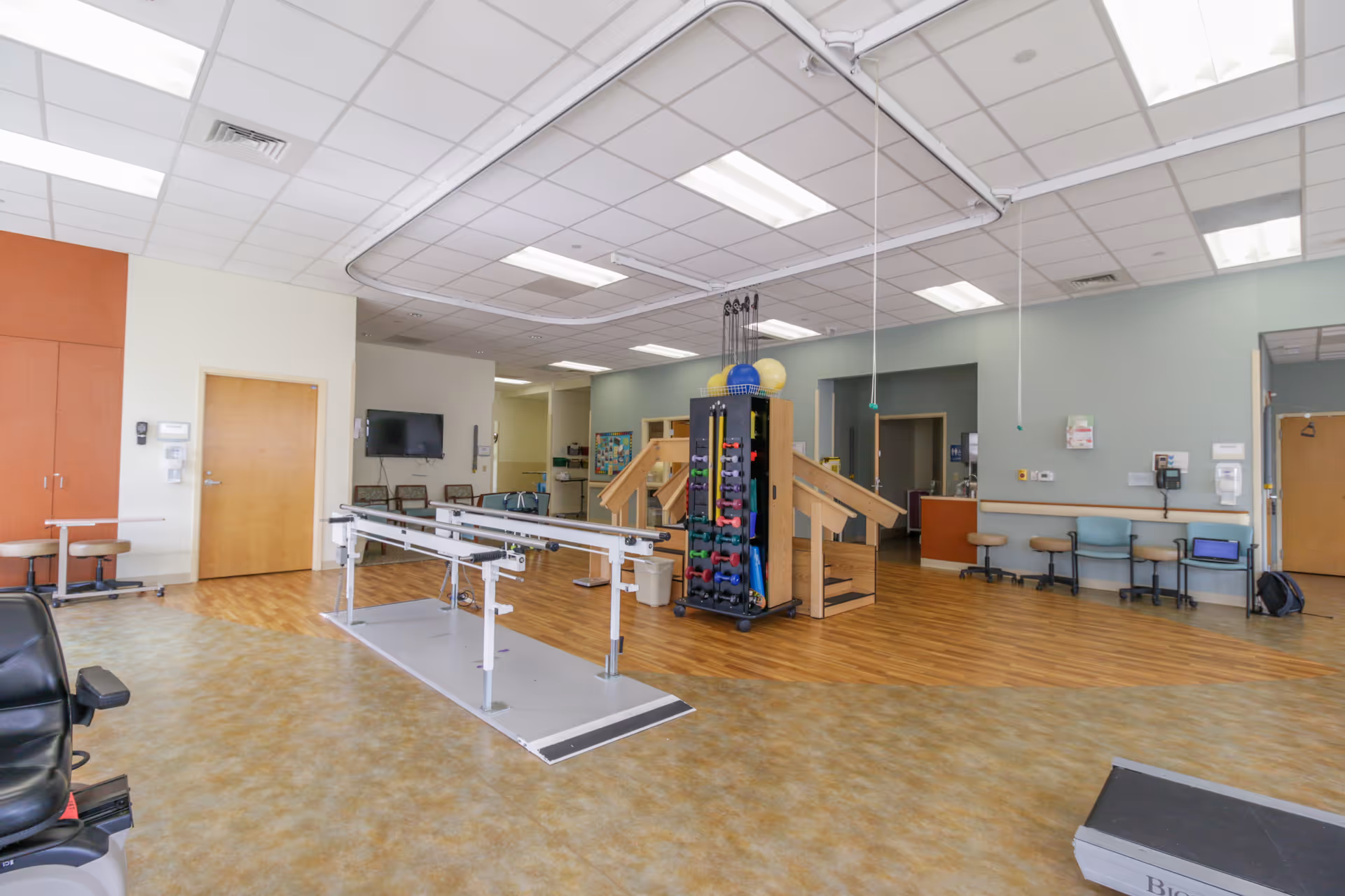 A spacious rehabilitation or physical therapy room with parallel bars for walking exercises, a rack holding various exercise equipment like dumbbells and therapy balls, wooden stairs for physical therapy, chairs along the walls, and a television mounted on the wall. The room has a combination of wood and vinyl flooring and a drop ceiling with fluorescent lighting.