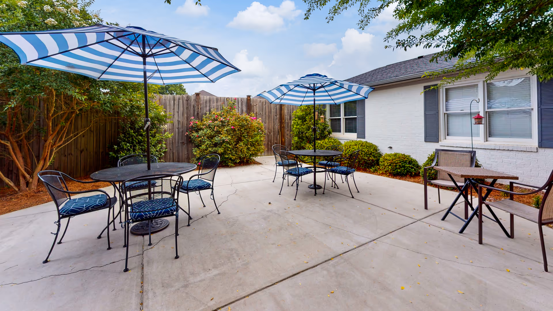 Outdoor patio area with two round metal tables, each with four chairs and blue and white striped umbrellas. There are also two additional chairs with a small table near a white brick building with windows and gray shutters. The patio is surrounded by greenery and a wooden fence.