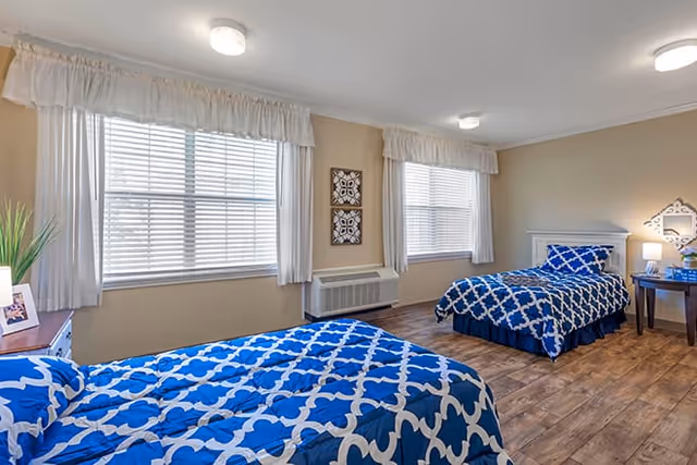 Bright shared bedroom with two twin beds in blue patterned bedding, wood-look floors, large windows with blinds and curtains, and small bedside tables.