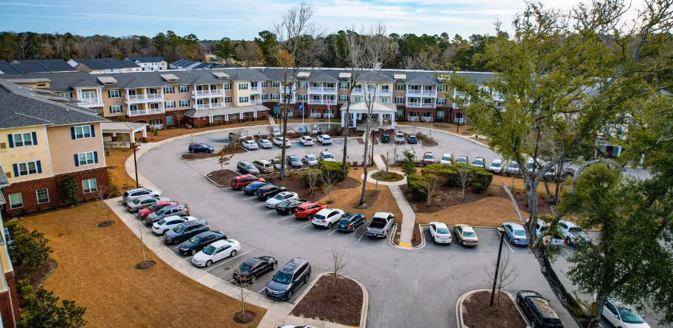 Aerial view of a senior living facility named Harmony at Wescott showing a large three-story building with multiple balconies, a circular driveway with parked cars, landscaped areas with trees and shrubs, and a clear sky above.