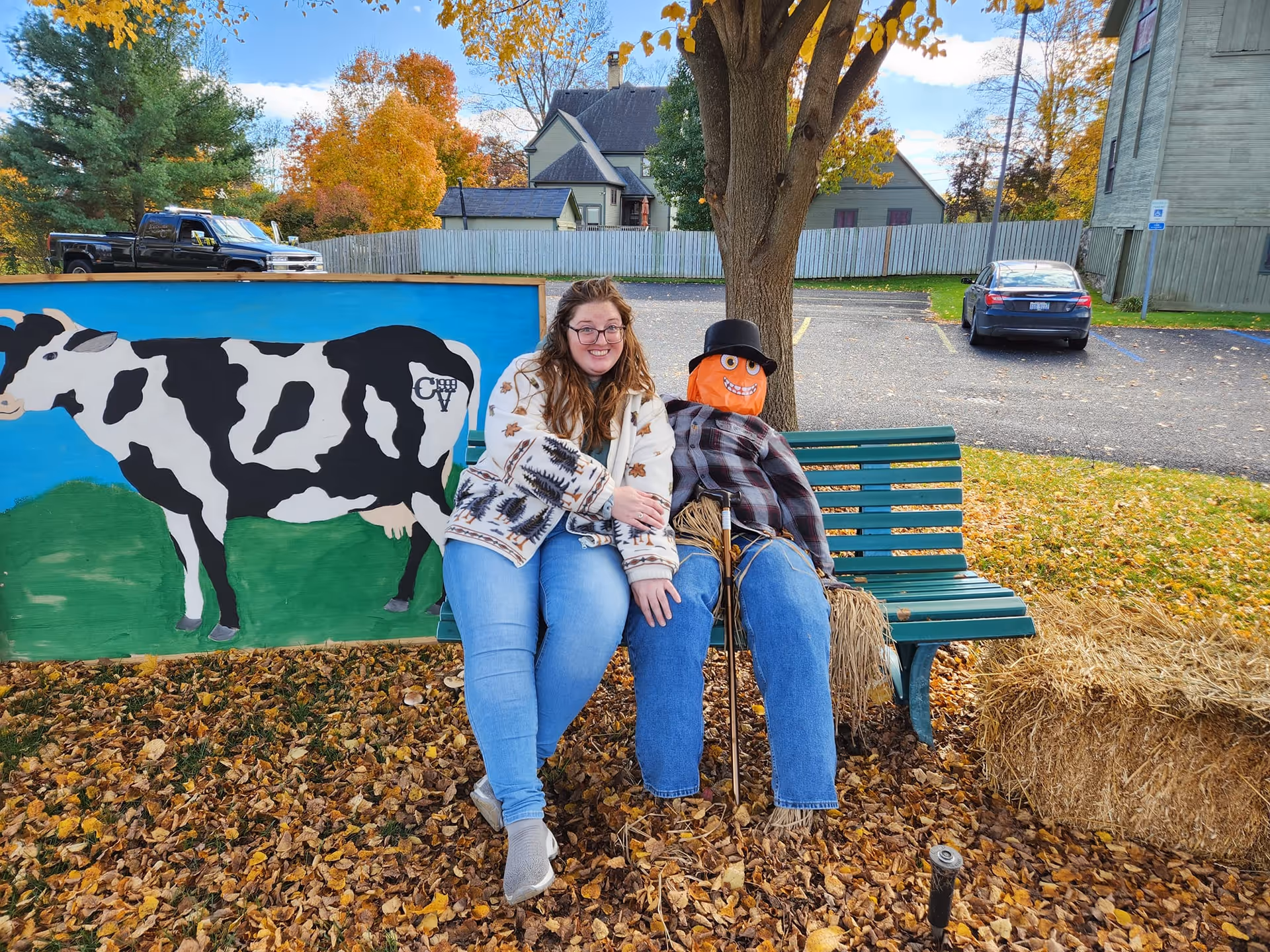 A woman with glasses and a patterned jacket sits on a green bench outdoors next to a scarecrow dressed in a plaid shirt, jeans, and a black hat. Behind them is a painted wooden board featuring a black and white cow with the letters 'CV' on its side. The ground is covered with fallen autumn leaves, and there are trees, parked cars, and houses in the background.