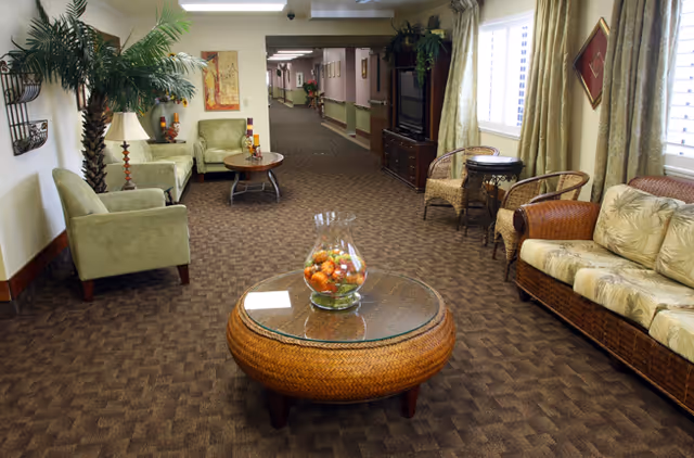 A carpeted senior living lounge with wicker and upholstered seating, a round glass-topped coffee table with decorative fruit, potted palms, and a long hallway in the background.