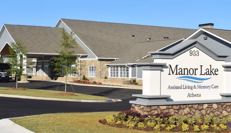 Exterior view of Manor Lake Assisted Living & Memory Care facility in Athens, showing the building with stone and siding facade, a driveway, small trees, and a large sign with the facility name and address number 933.