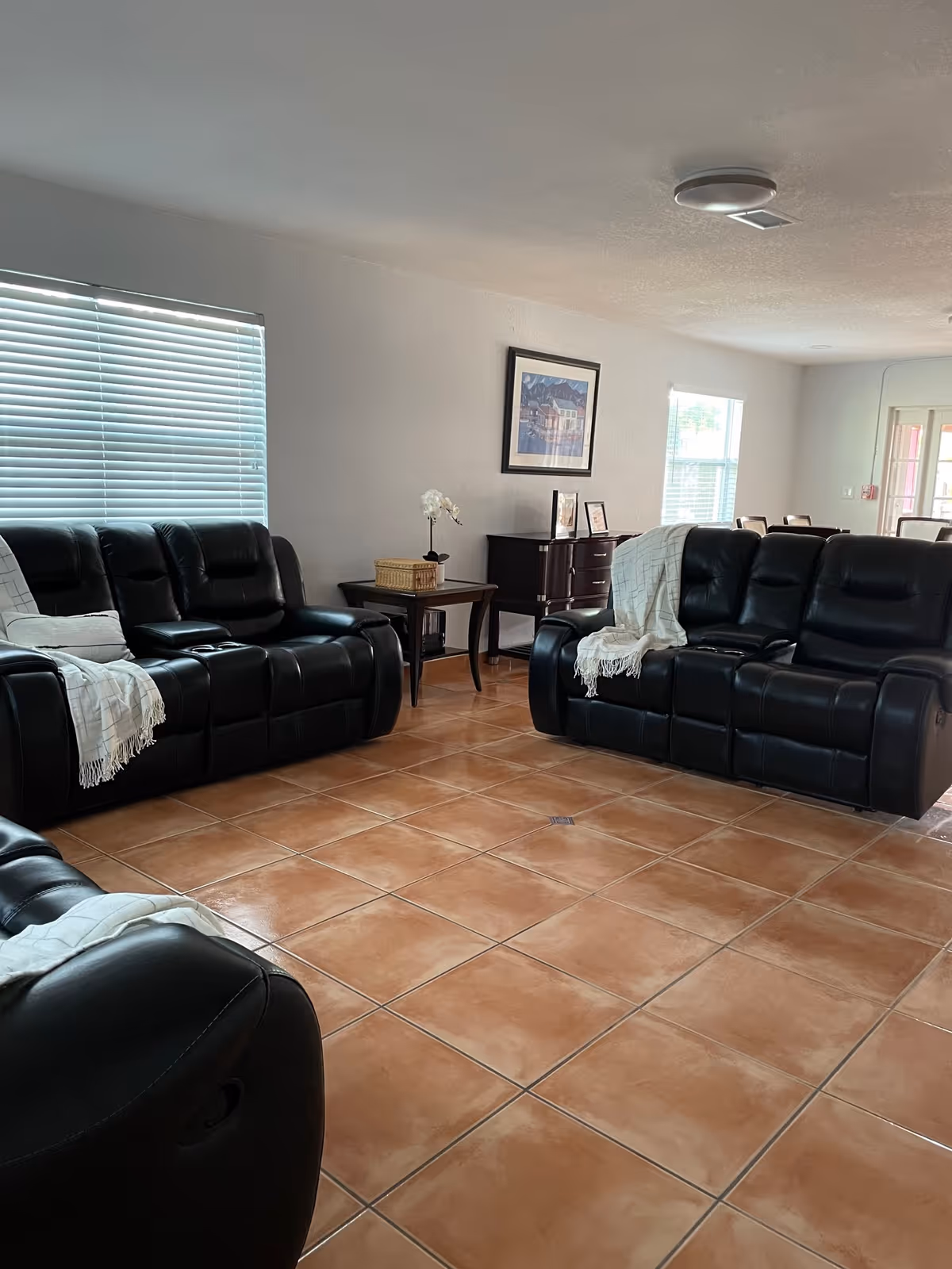 Spacious living room with black leather sofas, terracotta tile floor, side tables and framed art on the wall.
