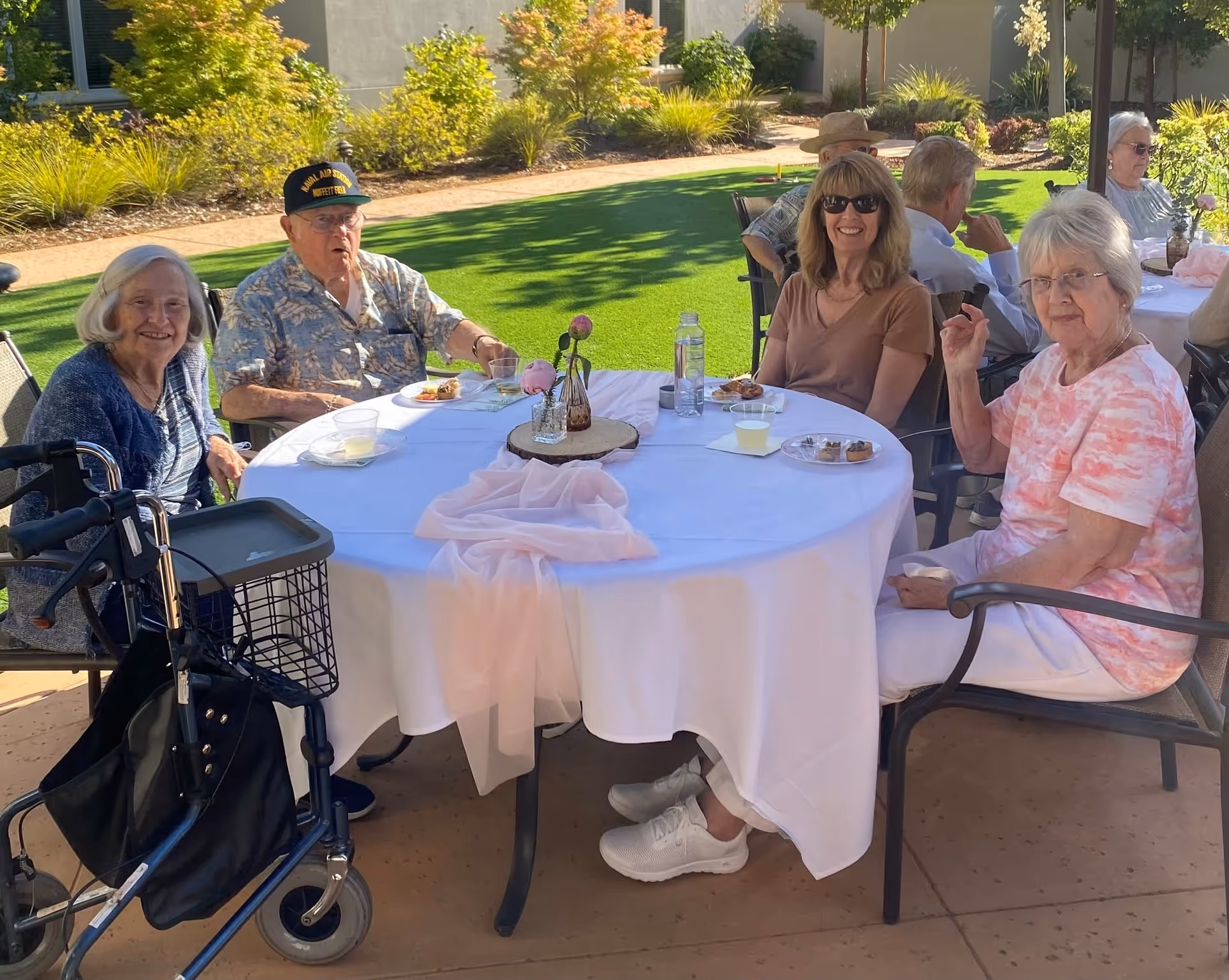 A group of elderly people sitting around a round table outdoors at Oakmont of Fair Oaks. The table is covered with a white tablecloth and has small plates with food and drinks. The setting is sunny with green grass and bushes in the background. Some individuals are smiling and appear to be enjoying the gathering.