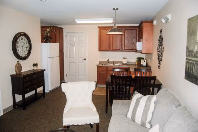 Interior view of a small assisted living apartment showing a living area with a gray couch and white armchair, a dining table with four chairs, and a kitchenette with wooden cabinets, a refrigerator, and a sink. The walls are light-colored and decorated with a clock, a wall art piece, and a decorative metal wall hanging.