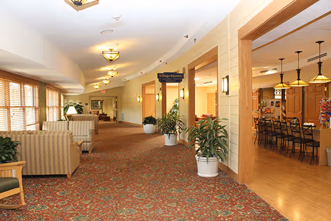 Carpeted corridor in a senior living facility with sofas and plants along the left and an open dining area visible to the right.